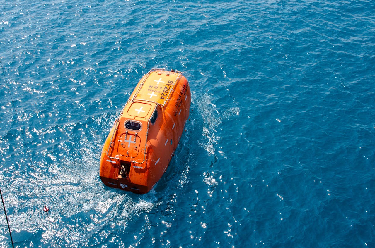 A bright orange lifeboat floating on the vivid blue ocean water, seen from above.