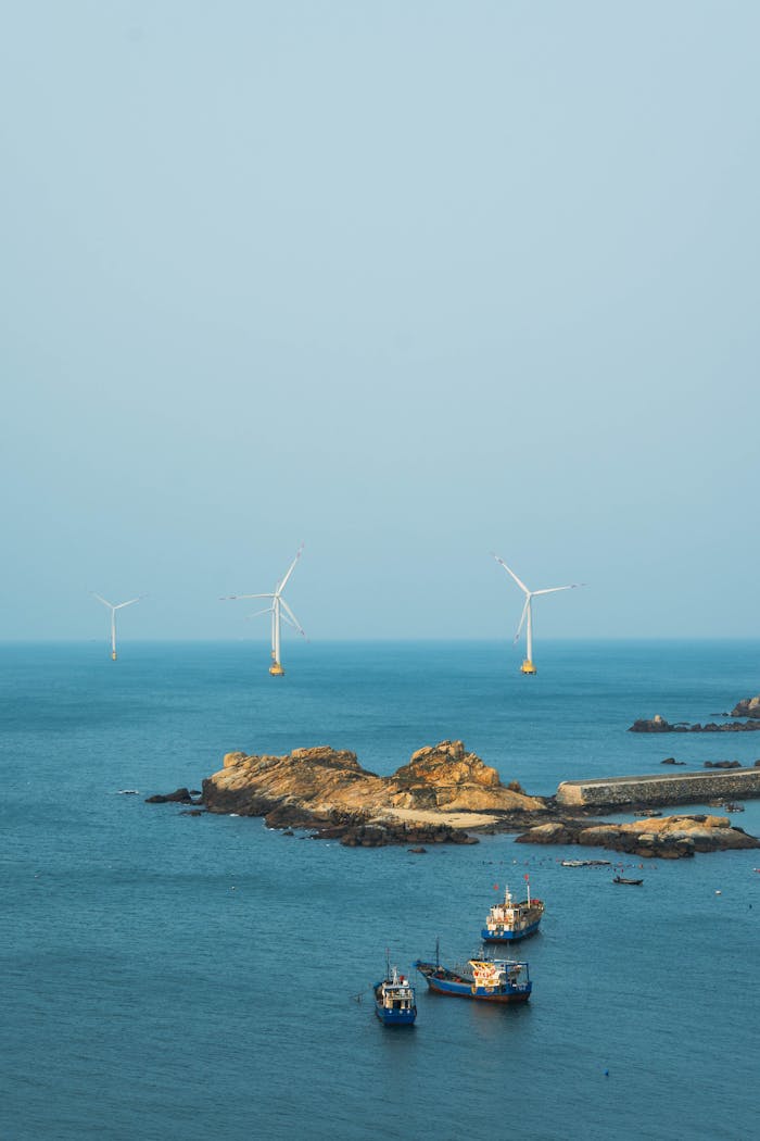 Serene image of wind turbines off a rocky coast with fishing boats in blue sea, showcasing renewable energy.